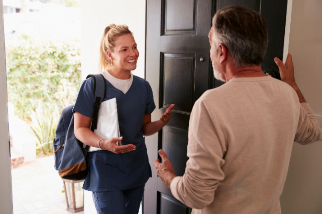 En el umbral de la puerta de una casa hay dos personas, en la parte interna de la casa hay un hombre y en la parte externa hay una mujer que por su vestimenta se puede inferir que es funcionaria del sector salud, ella tiene una cara amable y parece estar explicando algo. 