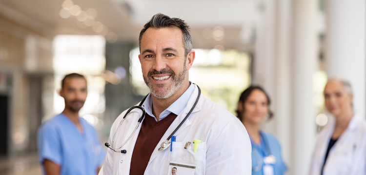 En un primer plano se ve a un médico sonriendo y en la parte de atrás se ven a otros profesionales de la salud mirando hacía el frente. 