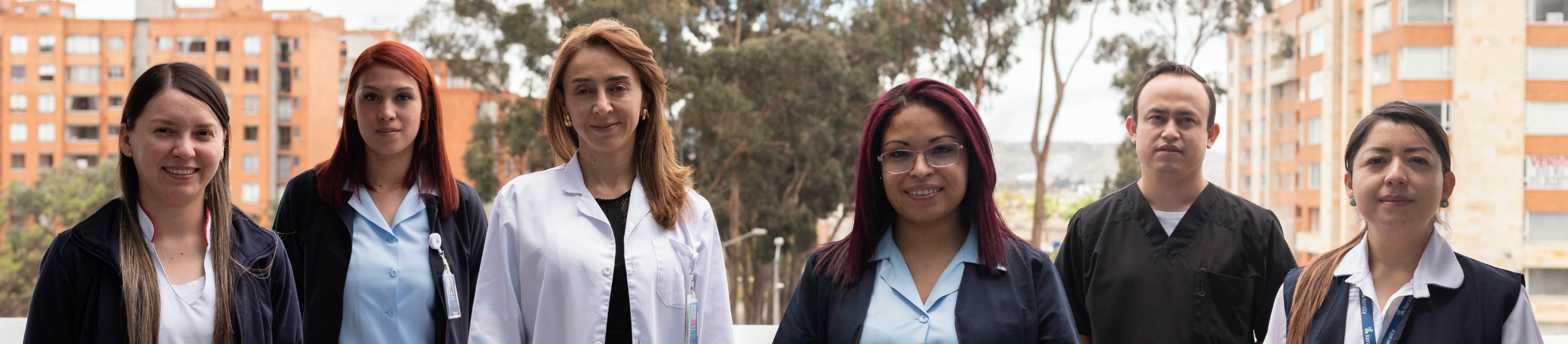 En un ambiente al aire libre en donde de fondo se pueden ver árboles y edificios se ve a 5 personas entre hombres y mujeres, profesionales de la salud y administrativos sonriendo y que se puede inferir que son colaboradores de la clínica. 