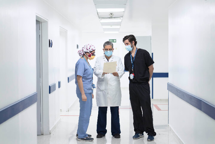 En un pasillo de una Clínica  se ven a dos doctores y una doctora revisando un documento, los tres están de pie. 