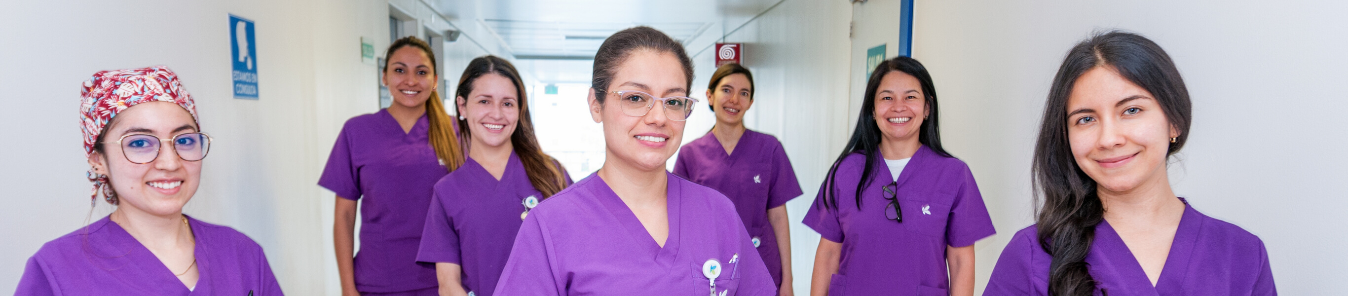 En un pasillo de una clínica se ven seis mujeres profesionales de la salud sonriendo. 