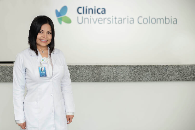 Se ve una mujer sonriendo con una pared de fondo que dice Clínica Universitaria de Colombia 